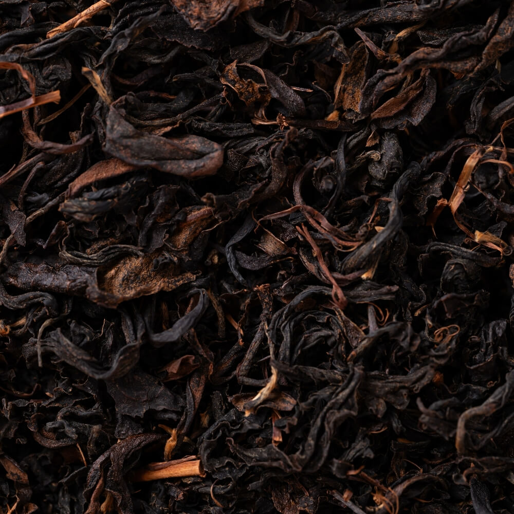 A close-up texture shot of dark, dried tea leaves, showing curled and twisted pieces in deep brown and black tones.