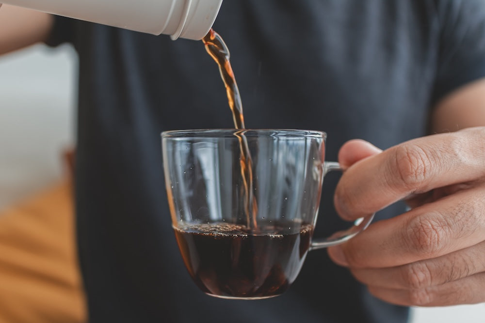 Coffee being poured from a white container into a clear glass mug held by a person wearing a dark gray t-shirt.