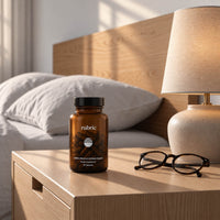 An amber supplement bottle labeled 'rubric' sits on a light wood bedside table next to a lamp and eyeglasses, with a bed in the background.