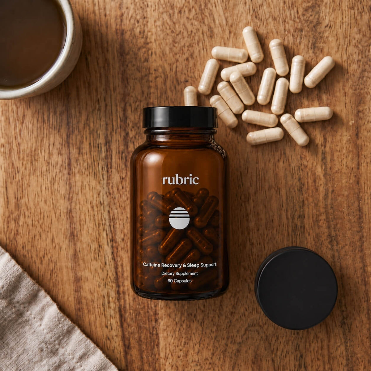 An amber glass supplement bottle labeled rubric, shown open with capsules spilled around it on a wooden surface, alongside a black cap and a cup of coffee.