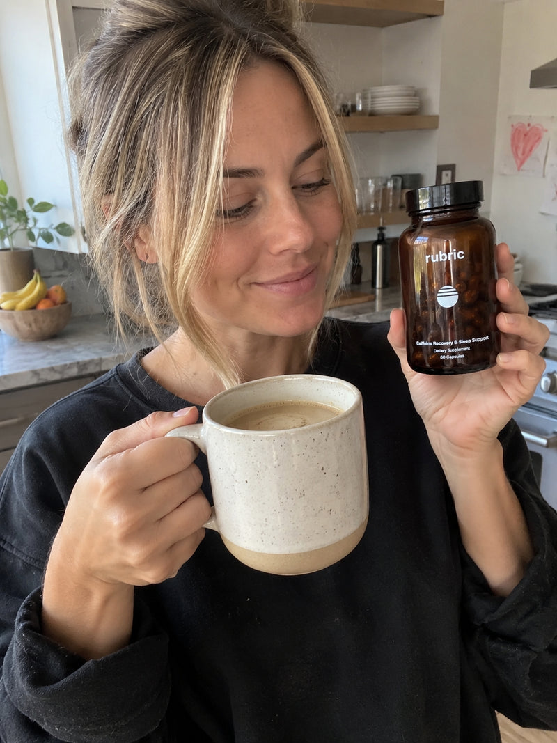 A woman in a black sweatshirt holds a ceramic mug and a brown amber bottle labeled 'rubic' in a bright kitchen setting.