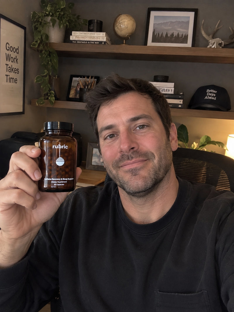 A man in a black crewneck sweatshirt holds up a brown amber glass bottle of Rubric brand dietary supplement capsules in front of a home office background with wooden shelves.