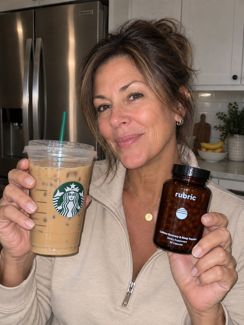 A woman wearing a beige sweater, standing in her kitchen holding a Starbucks coffee and the Rubric bottle